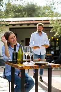 Federico e Giada nel dehors tra gli ulivi del wine bar di Petricor, meta perfetta per amanti dell'oleoturimo: lei è seduta a un tavolo con del vino davanti e lui sta portando dei piattini da mangiare